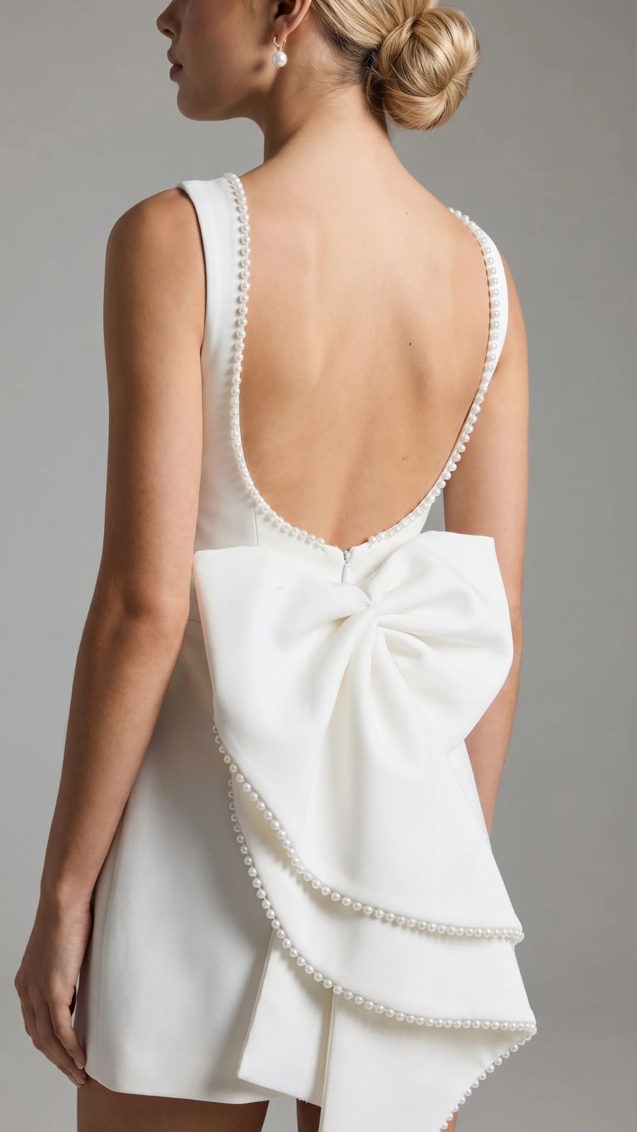 Back view of a woman with an updo hairstyle, wearing a white backless mini dress with a large pearl-edged bow and pearl details on the open back.