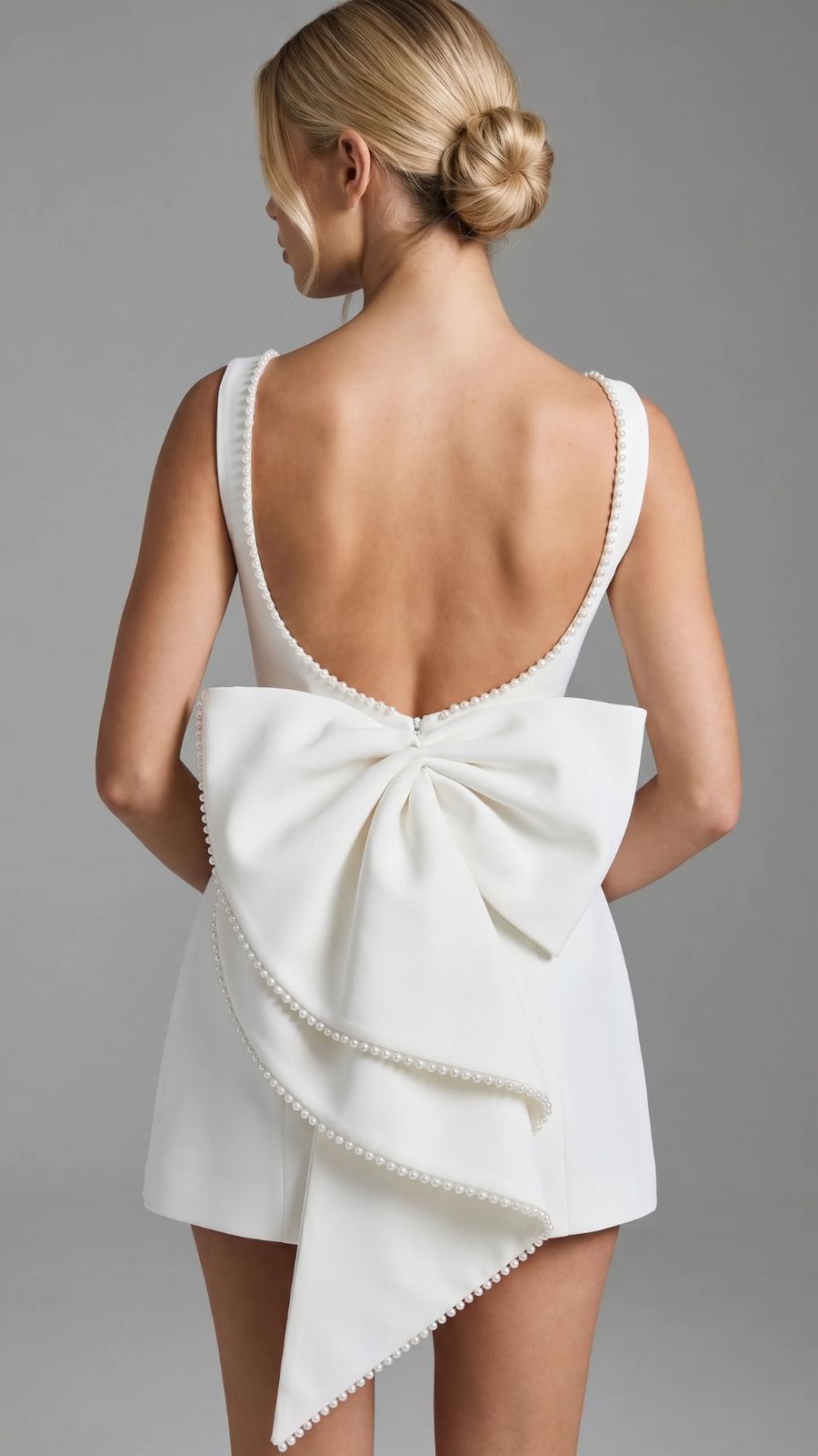 Rear view of a woman in a white mini dress, showing the deep scoop back adorned with pearls and a prominent, pearl-trimmed bow cascading down the skirt.