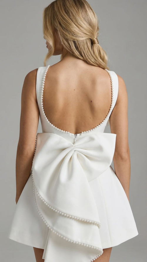 Back view of a woman wearing a white mini dress with a large pearl-trimmed bow and a backless design with pearl accents along the neckline.