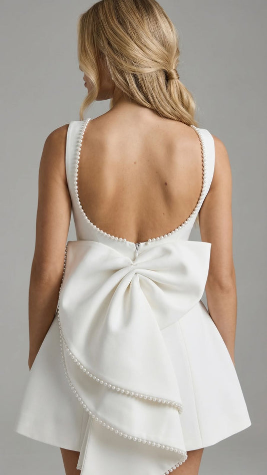 Back view of a woman wearing a white mini dress with a large pearl-trimmed bow and a backless design with pearl accents along the neckline.