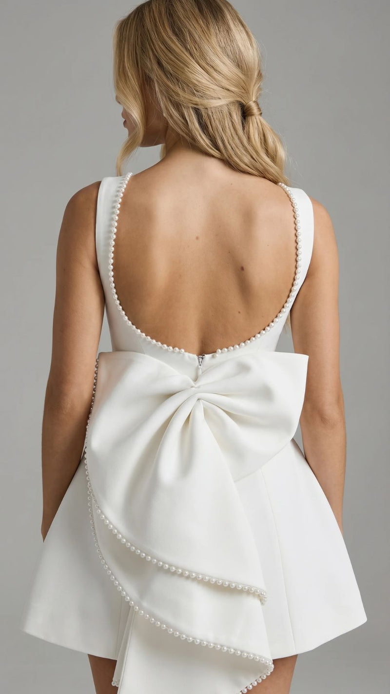 Back view of a woman wearing a white mini dress with a large pearl-trimmed bow and a backless design with pearl accents along the neckline.