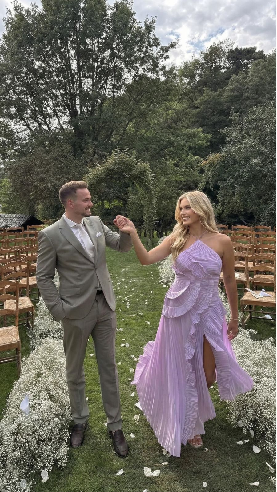 Blonde woman in a lavender Nathalie ruffle maxi dress with a thigh-high slit, holding hands with a man in a suit, walking on grass.
